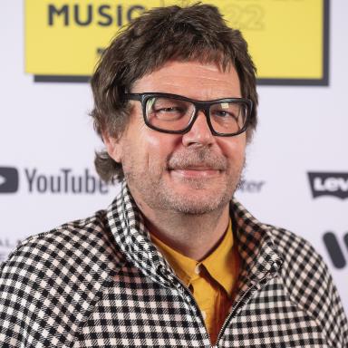Man with black framed glasses wearing black & white check shirt, in front of a background board saying Youth Music Awards 2022