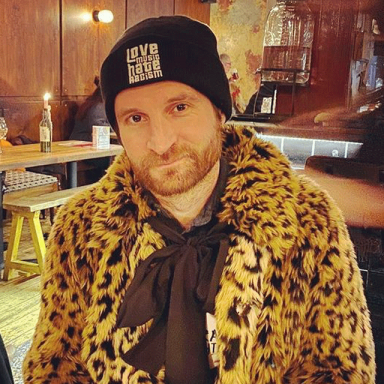Paul wearing a fluffy leopard-print coat tied with a bow, and a Love Music Hat Racism beanie