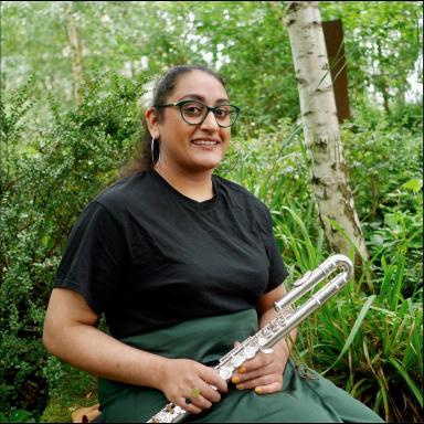 Diljeet Kaur Bhachu holds a flute and poses seated against greenery
