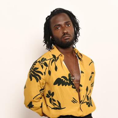 kwabs wears a yellow shirt with black floral prints and stands with his hands behind his back, leaning against a white wall