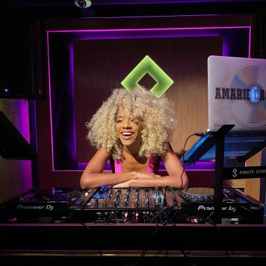 Amarie has curly hair wears a pink top and leans on a DJ deck