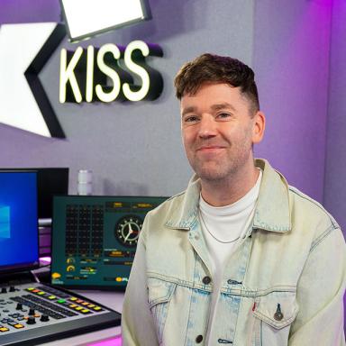 nathan thomson sits and smiles in the KISS radio office. he wears a white t shirt and a light blue denim jacket