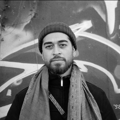 a man wears a scarf and beanie and poses against a graffiti wall. the photo is in black and white