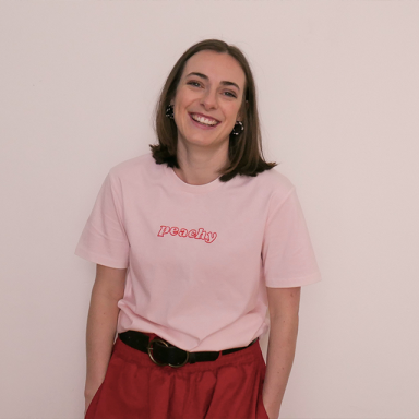 a woman with shoulder length, straight brown stands against a cream wall with her hands in her pockets. she is smiling and wearing a pink t-shirt with the word 'peachy' on it.