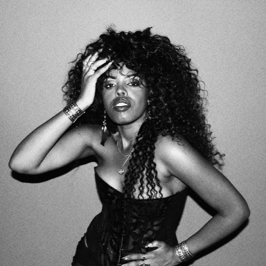 a woman with curly hair wears a strapless top and poses for the camera with one hand on her hip and one hand on her forehead