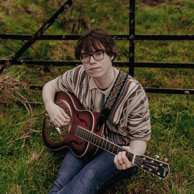 niall mcdowell sits on the grass next to a fence and holds a guitar. they have brown hair and wear glasses