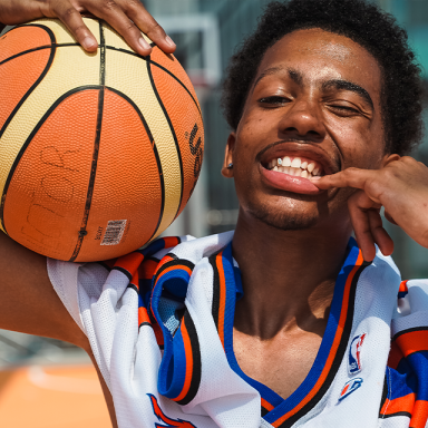 a young man holds a basketball on his shoulder and points to his teeth with a finger