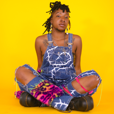 TU3SDAY wears dungarees and sits with their legs crossed. the background is bright yellow