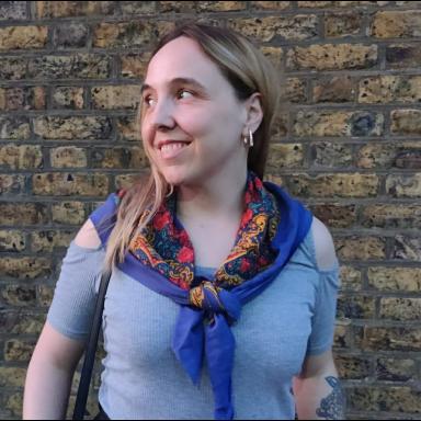 a woman with blonde hair stands in front if a brick wall and looks to the side, smiling. she wears a blue top and a dark blue patterned scarf