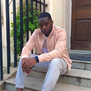Ife sits on stairs outside of a house. he is resting his elbows on his knees. he is wearing a pink overshirt and light blue jeans