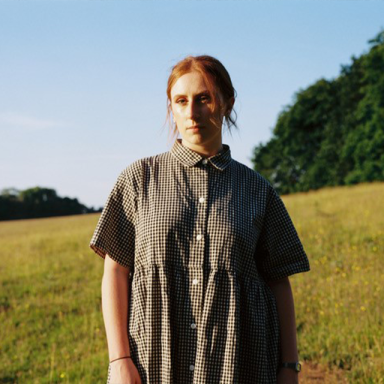 malan wears a plaid oversized shirt and stands in a field. her auburn hair is tied back.