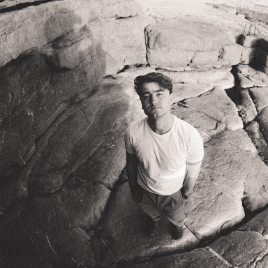tom auton poses on some rocks. he wears a white t shirt and has his hands in his pockets