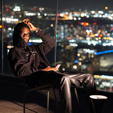 ambrose hill sits on a chair in a black hoodie and trousers. it is night and the skyline glows behind them.