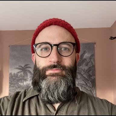 a man with a grey beard wears black glasses and a red beanie