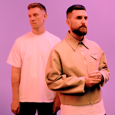 two men stand facing away from each other against a pink background