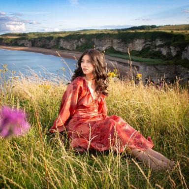 a woman in a floral dress has long brown wavy hair. she sits on the grass on a cliff, the water behind her
