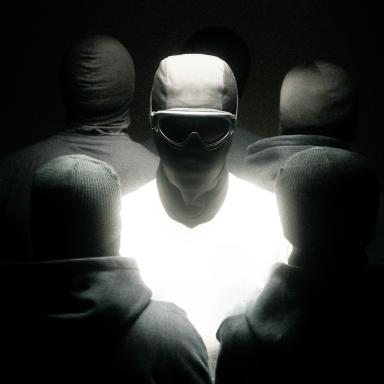 a person has their face covered by a mask but their body is illuminated white. they are surrounded by several hooded figures with their backs turned