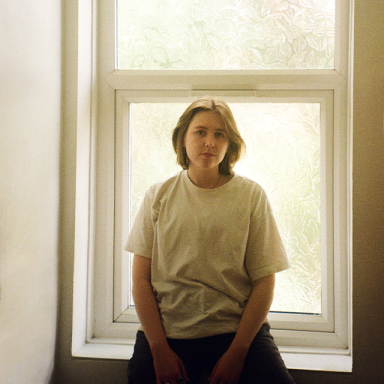 a woman with short hair wears a white t-shirt and sits on a windowsill inside