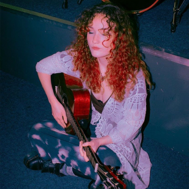 a woman with curly red hair sits on the floor and plays guitar