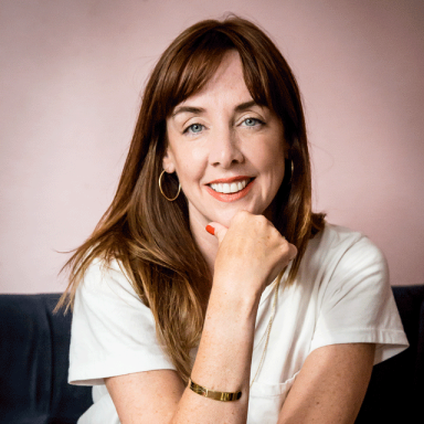 a woman with brown hair and bangs smiles and rests her chin on her fist. she wears a white t-shirt and is seated, leaning forward