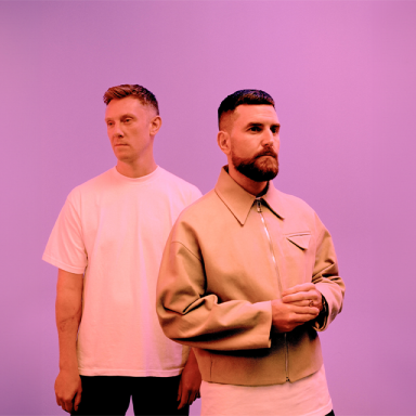 two men stand against a pink background and pose