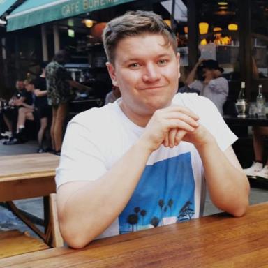 a man with cropped brown hair and a white t shirt rests his elbows on a wooden table