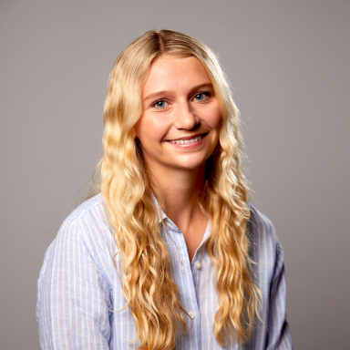 a woman smiles, wears a blue pinstripe shirt and has long wavy blonde hair