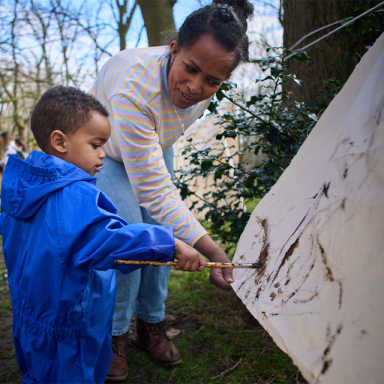 a boy paints outside on a canvas with help from an adult