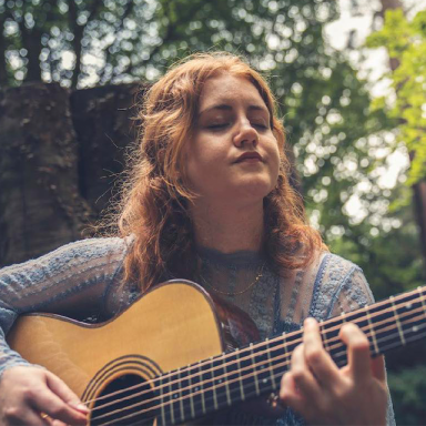 a woman with auburn hair closes her eyes and plays an acoustic guitar