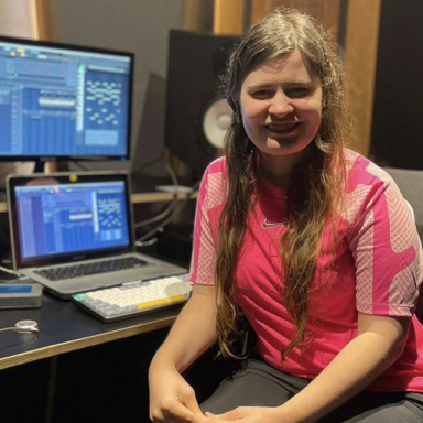 a woman with brown hair smiles and wears a pink t shirt in front of some computers with music software