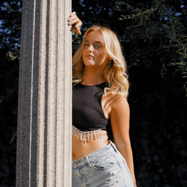 a woman with blonde hair, black crop top and blue jeans leans against a column