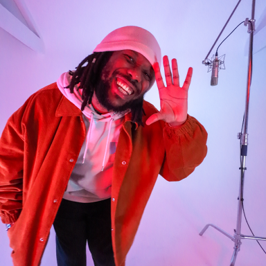 tee peters smiles, raises his palm and wears a red hoodie and white beanie